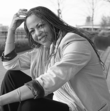 Poet Samiya Bashir poses in a lavender blazer on a roof, sitting with her left arm around her knee and right hand touching the top of her head